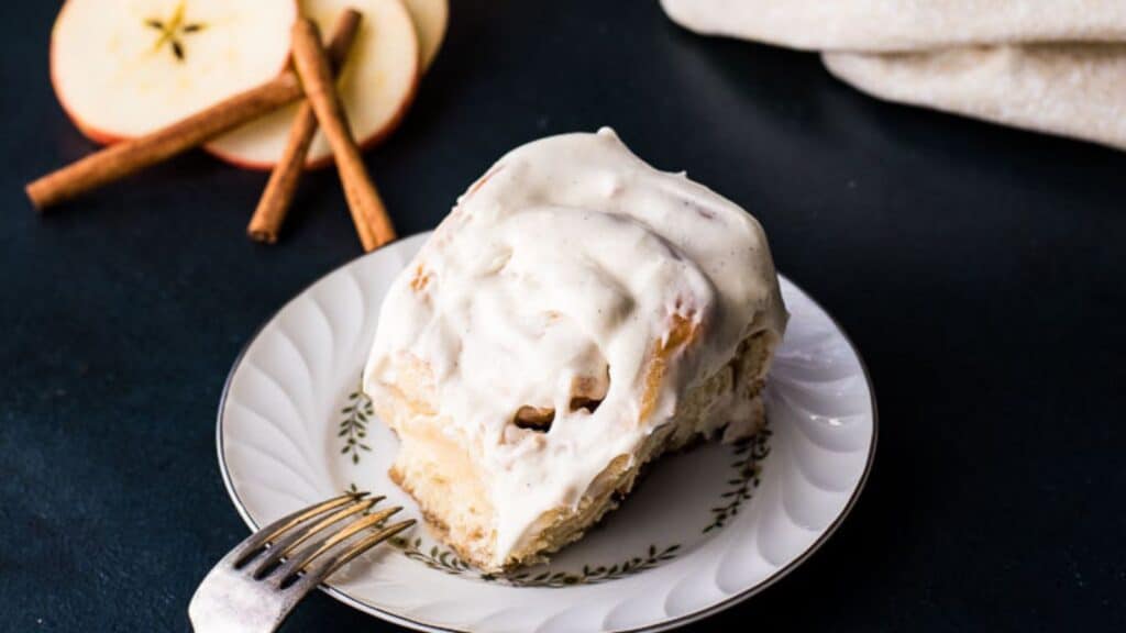 A slice of cinnamon apple cake with cream topping on a plate with a fork, accompanied by apple slices and cinnamon sticks.