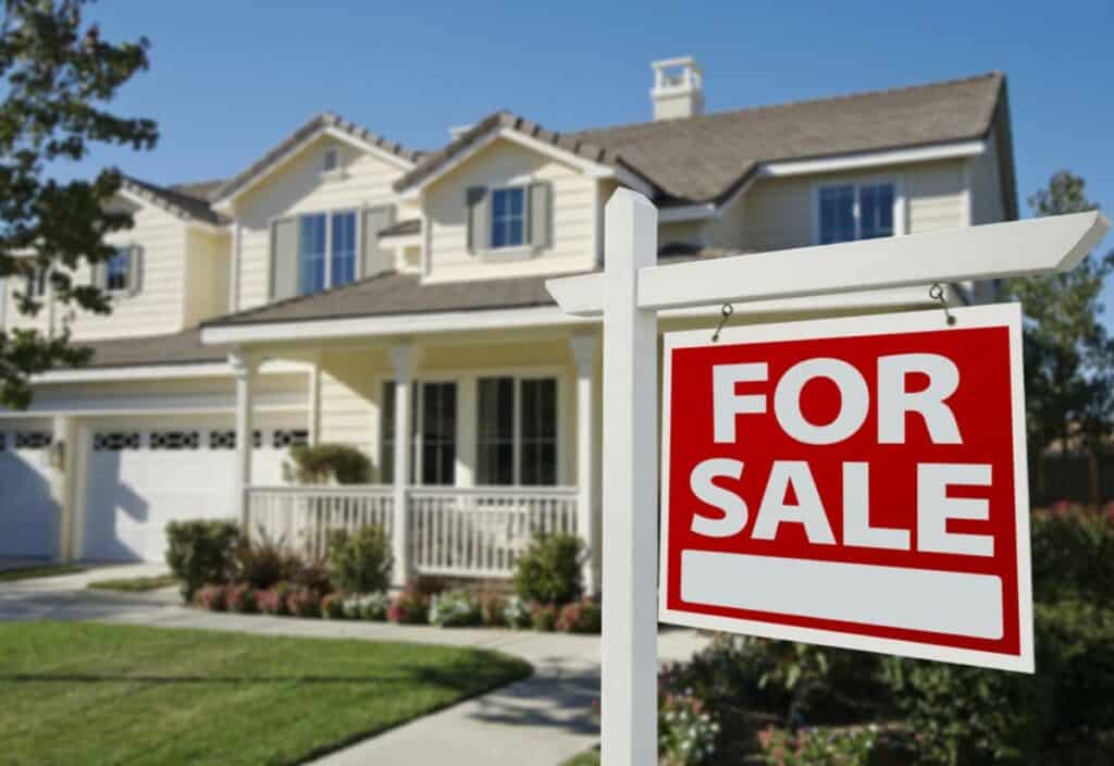 A "for sale" sign in front of a large, two-story suburban house with a landscaped yard under a clear sky.