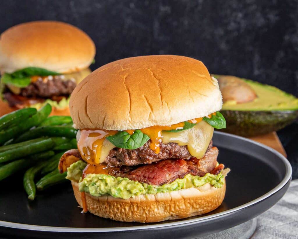 A gourmet burger with avocado, bacon, and melted cheese on a black plate, served with green beans.