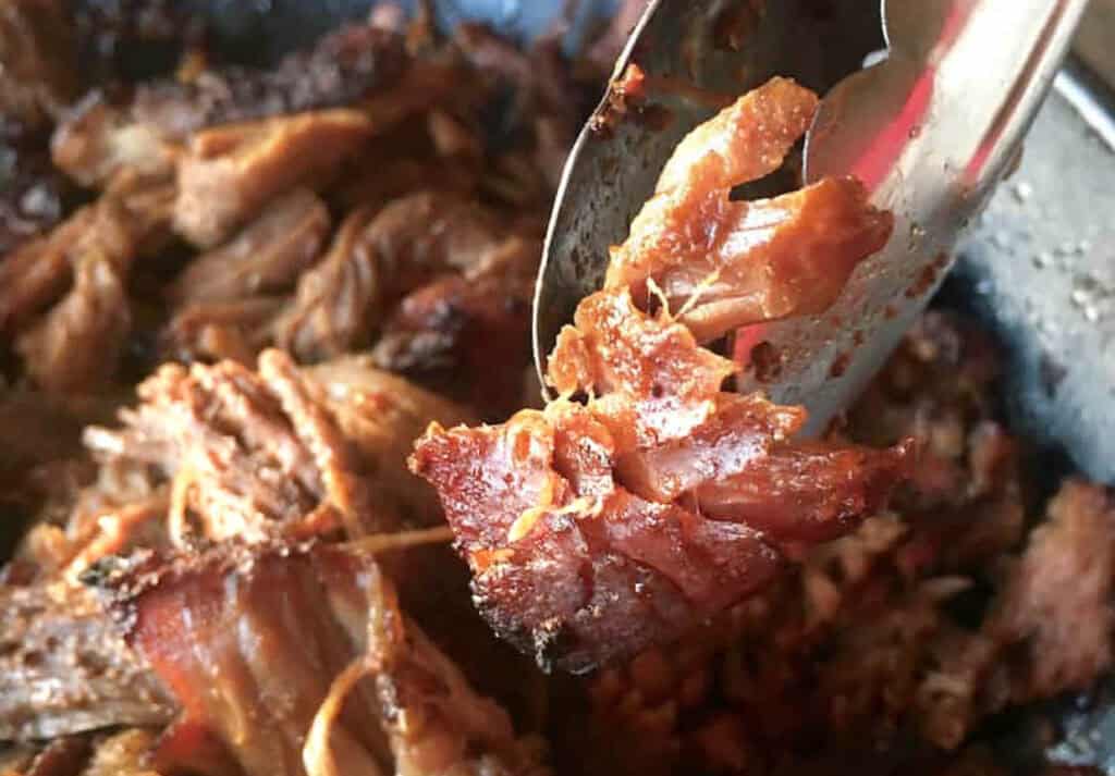 A close-up of shredded slow-cooked pork being served with tongs, highlighting the tender, juicy texture of the meat.