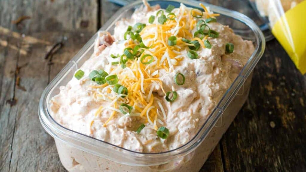 A container of creamy dip topped with shredded cheese and chopped green onions on a wooden surface.