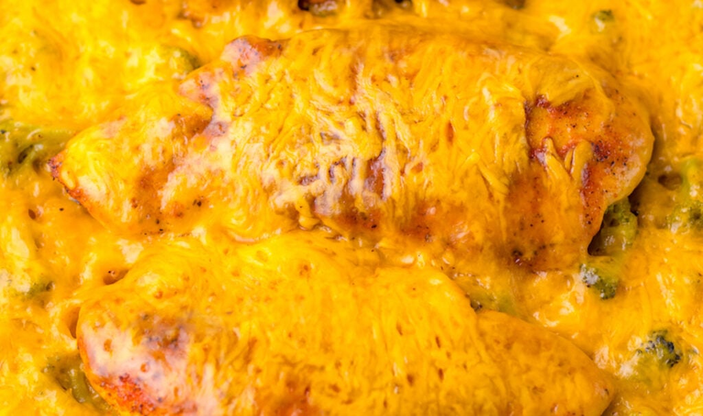 Close-up of a cheesy chicken enchilada dish with melted cheese on top, showing slight browning and visible textures of the sauce and fillings.