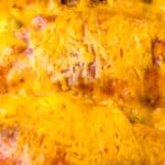 Close-up of a cheesy chicken enchilada dish with melted cheese on top, showing slight browning and visible textures of the sauce and fillings.