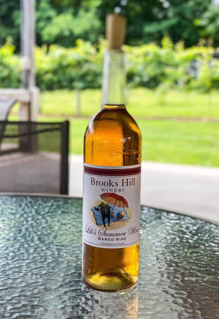 A bottle of brooks hill winery mango wine labeled "girls summer wine" on a glass table, with a blurred green garden background.