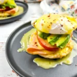 Eggs benedict with ham and avocado on an English muffin served on a black plate.