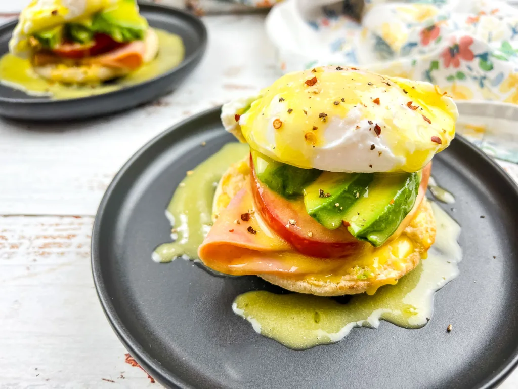 Eggs benedict with ham and avocado on an English muffin served on a black plate.