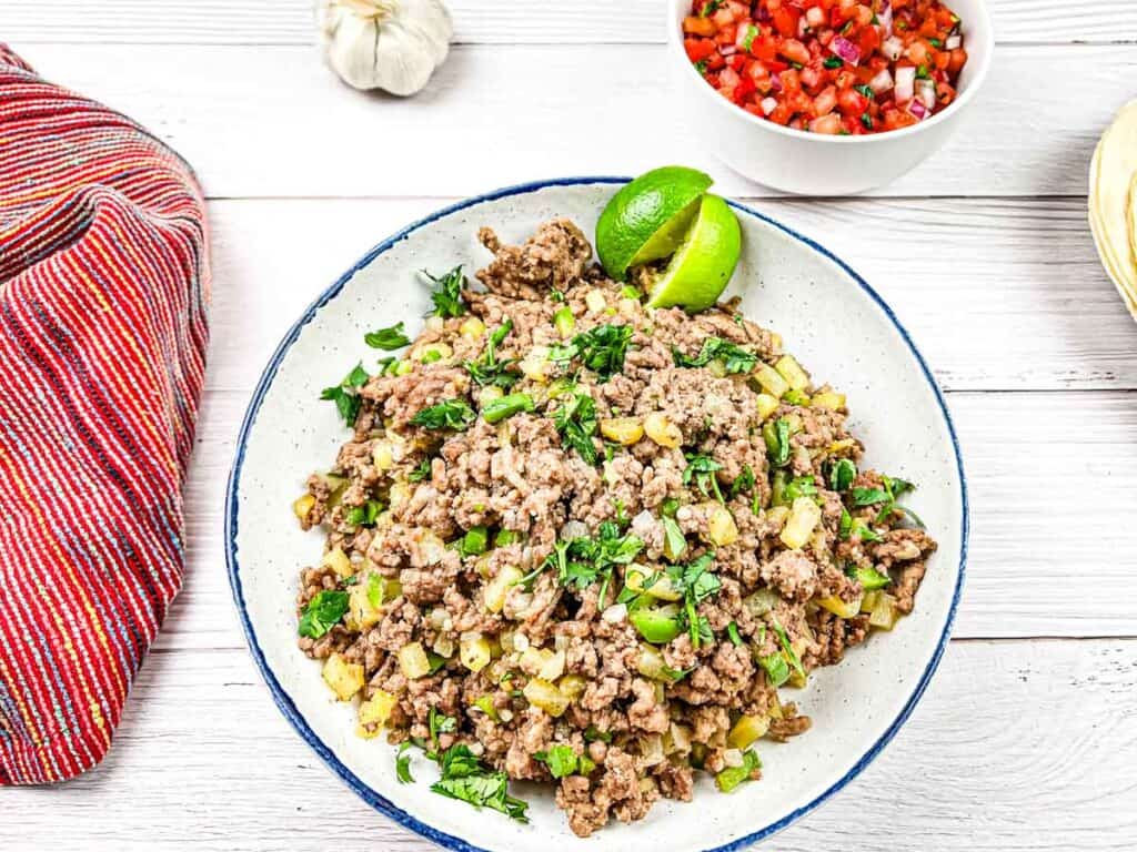 A plate of ground meat with diced potatoes and herbs, garnished with lime wedges, beside a bowl of salsa and garlic cloves on a wooden table.