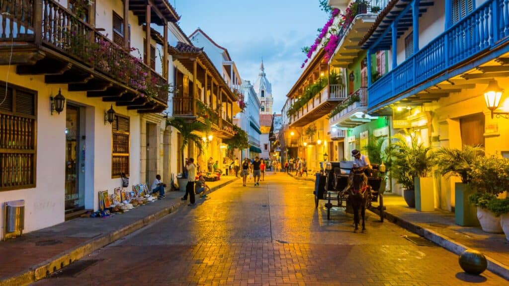 A vibrant street scene at dusk in cartagena, colombia, showing a horse-drawn carriage, pedestrians, and colonial buildings with balconies adorned with flowers.