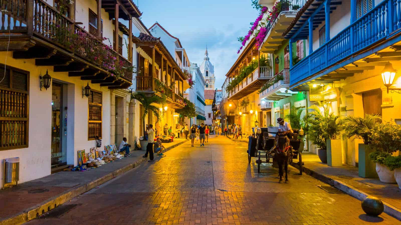 A vibrant street scene at dusk in cartagena, colombia, showing a horse-drawn carriage, pedestrians, and colonial buildings with balconies adorned with flowers.