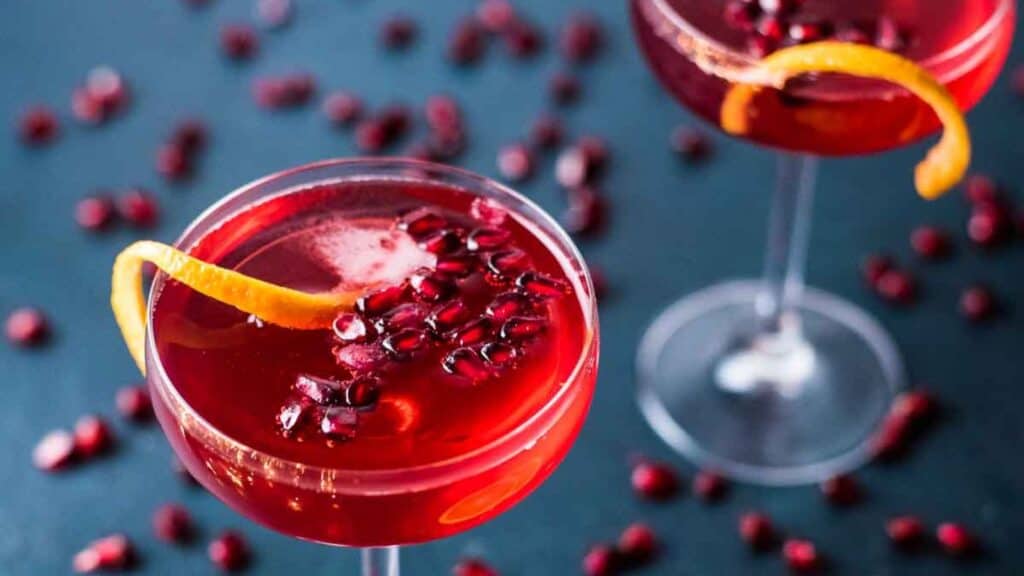 Two glasses of red cocktail garnished with orange peel and pomegranate seeds on a dark blue surface.