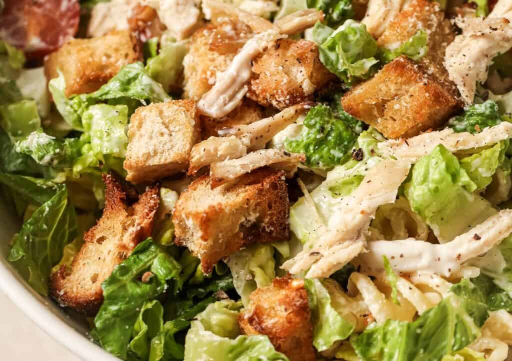 Close-up view of a caesar salad with croutons, grated cheese, and pieces of chicken, served in a white bowl.