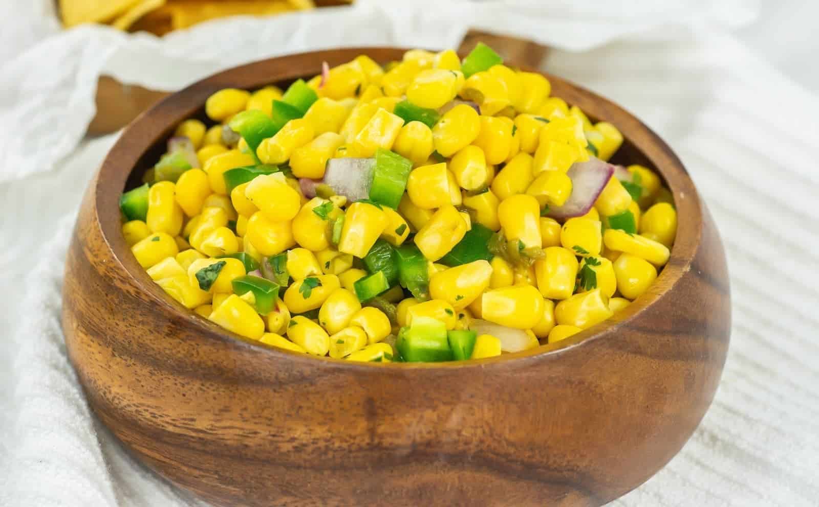 A bowl of corn salsa with diced green peppers and red onions, served in a wooden bowl on a white cloth.