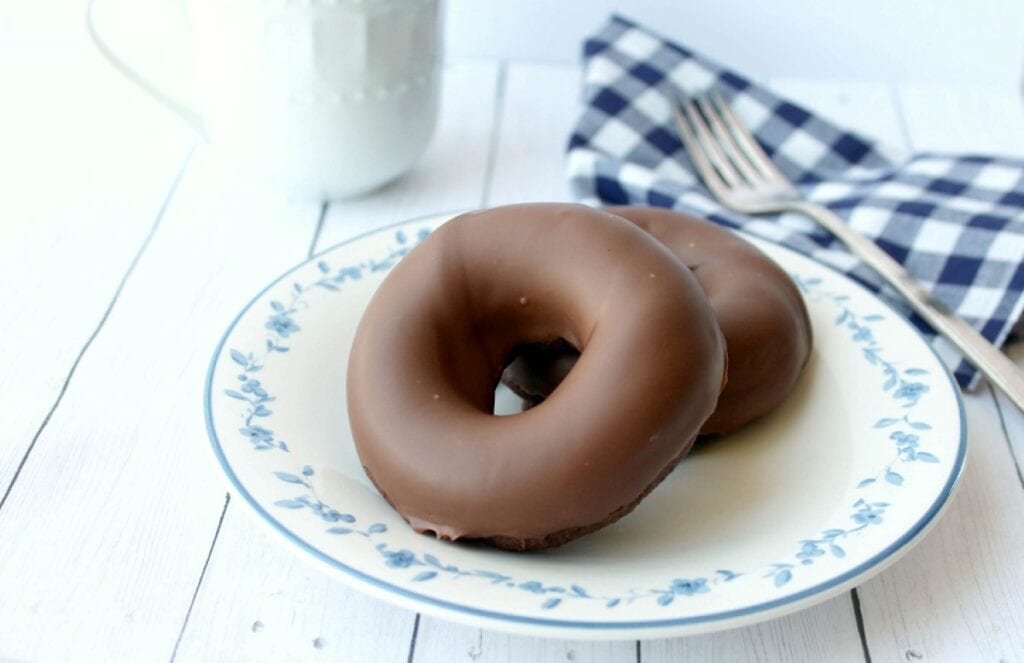 Two chocolate glazed donuts on a white and blue patterned plate with a fork and a napkin on the side.