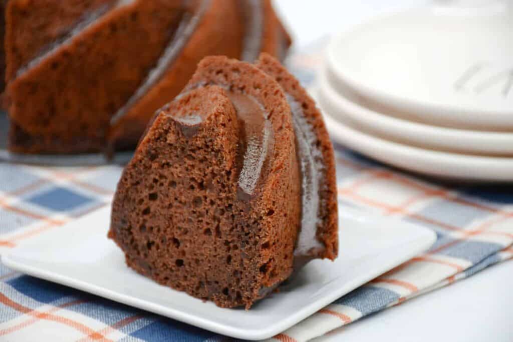 A slice of chocolate bundt cake on a white plate with the rest of the cake in the background.