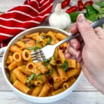 A hand holding a fork over a bowl of Fornaio Rigatoni Alla Vodka on a wooden table.