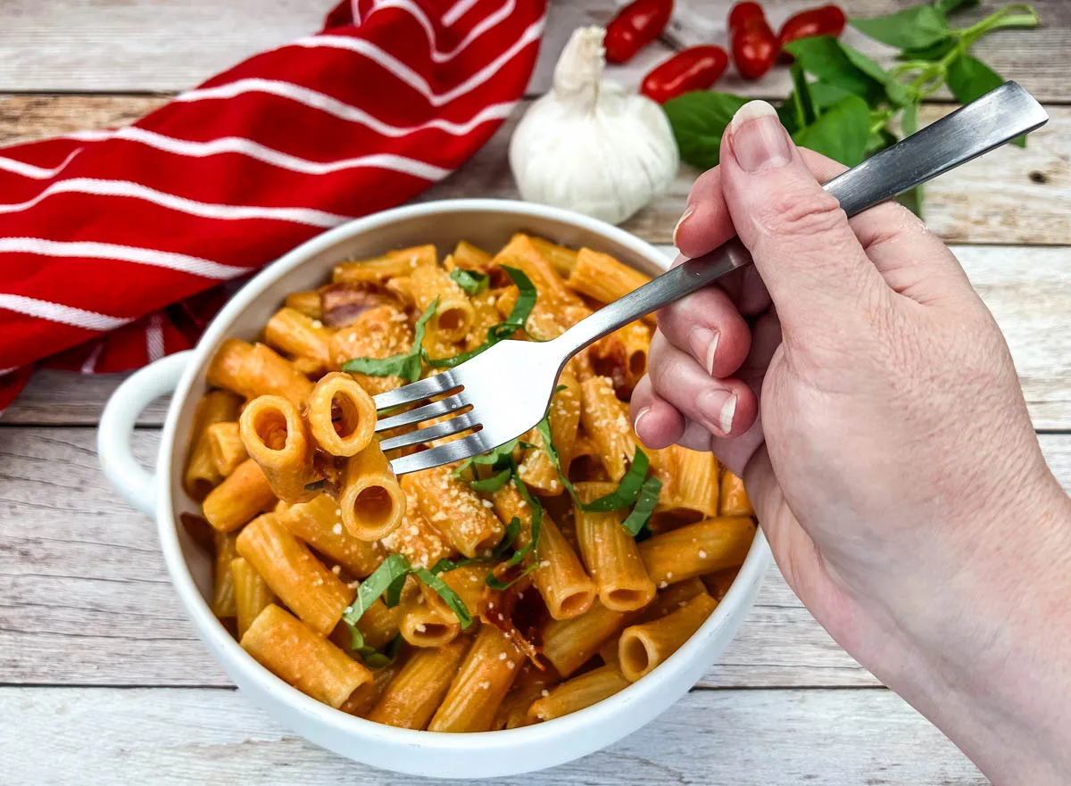 A hand holding a fork over a bowl of Fornaio Rigatoni Alla Vodka on a wooden table.