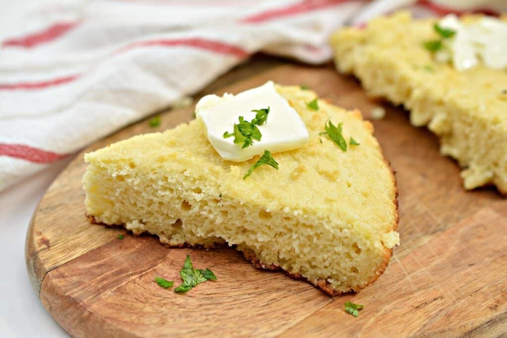 Slice of cornbread with butter on top on a wooden board, garnished with parsley, with a striped towel in the background.