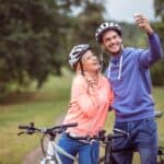 A couple takes a selfie with a smartphone, both wearing helmets and standing next to bicycles in a green park.