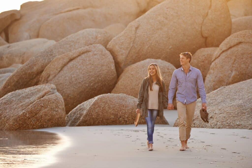 A young couple walking hand in hand on a sandy beach, surrounded by large boulders, during sunset.