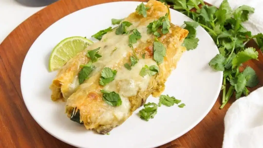 A plate of cheesy enchiladas garnished with fresh cilantro and a slice of lime.