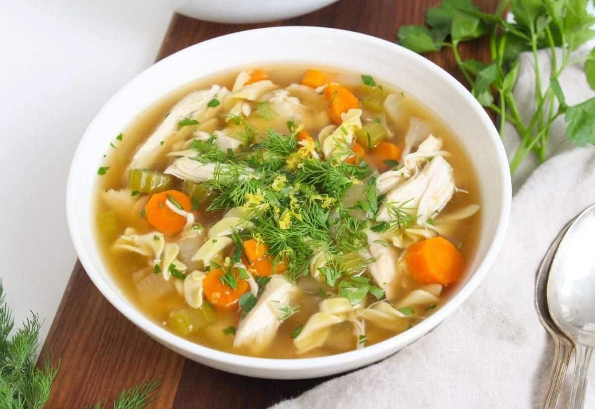 A white bowl with chicken noodle soup topped with fresh herbs.