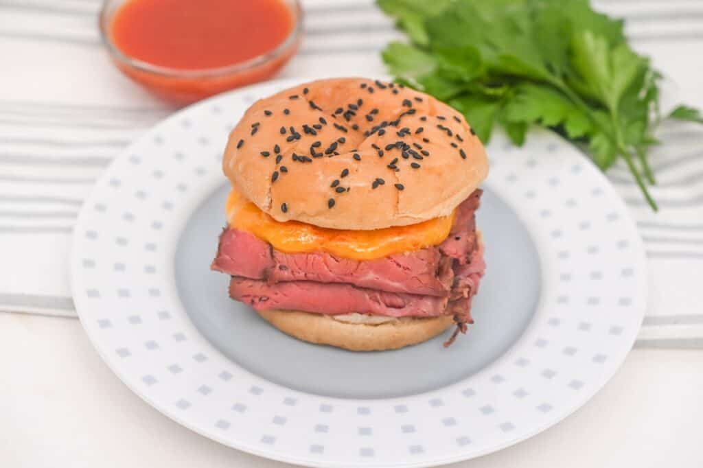 A roast beef sandwich with cheddar cheese on a sesame bun, served on a plate with a side of sauce and fresh greens.