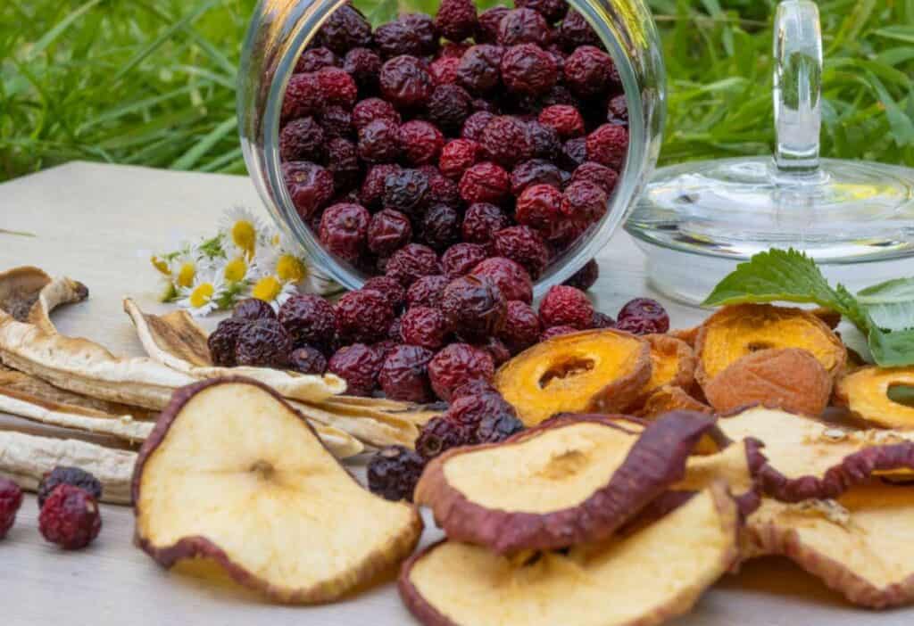 Glass jar tipped over, spilling dried cherries onto a wooden table, surrounded by assorted dried fruit slices and fresh raspberries on a grassy background.