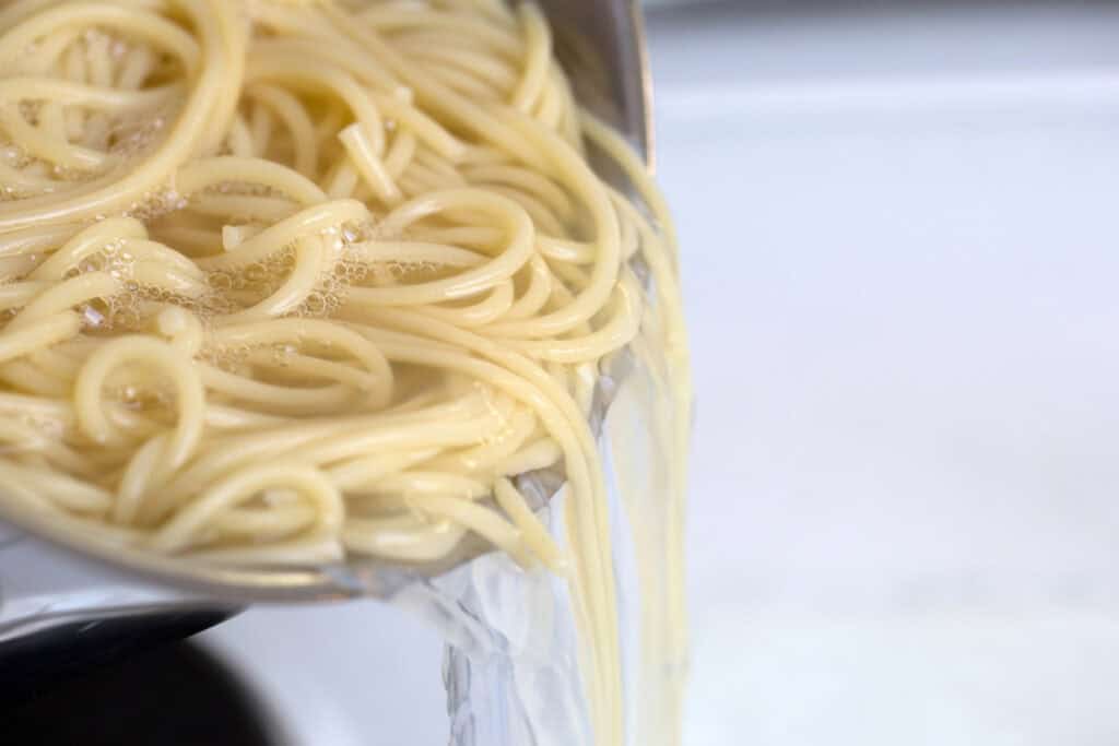 Cooked spaghetti noodles draining in a metal colander over a sink, with steam rising.