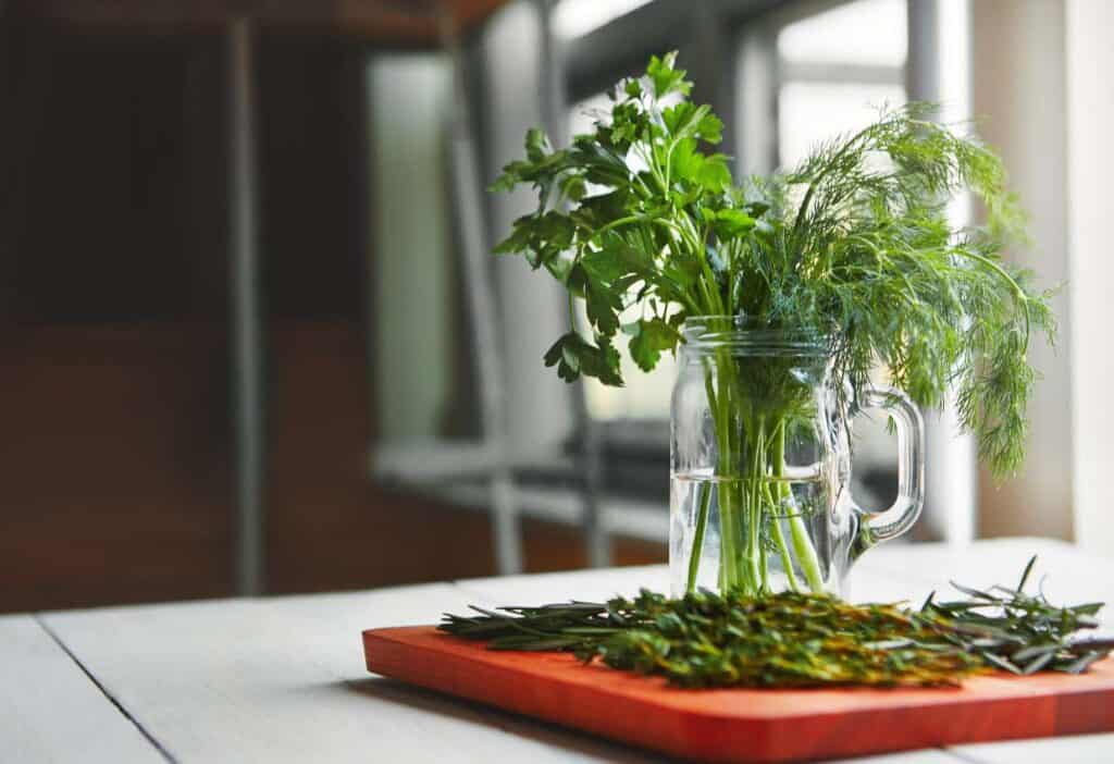 A glass jar with fresh parsley and dill on a wooden board with rosemary besides it.