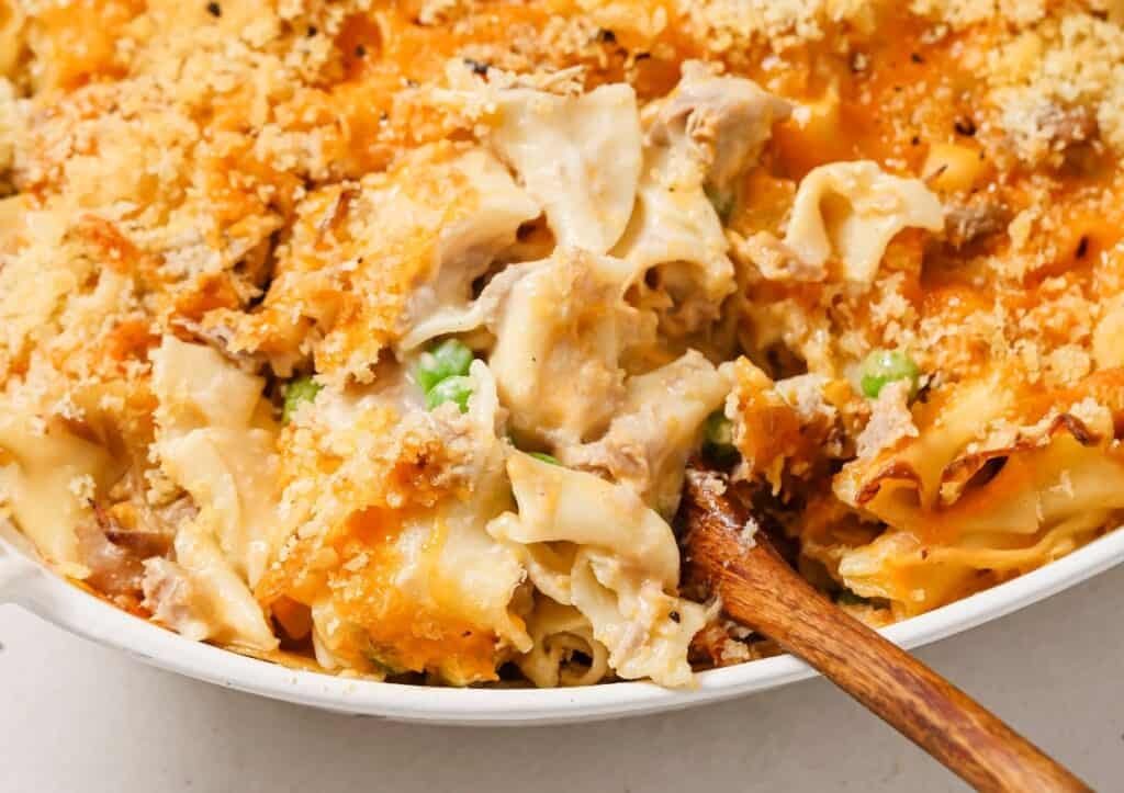 A creamy tuna noodle casserole with peas and a crispy breadcrumb topping in a white dish with a wooden spoon.