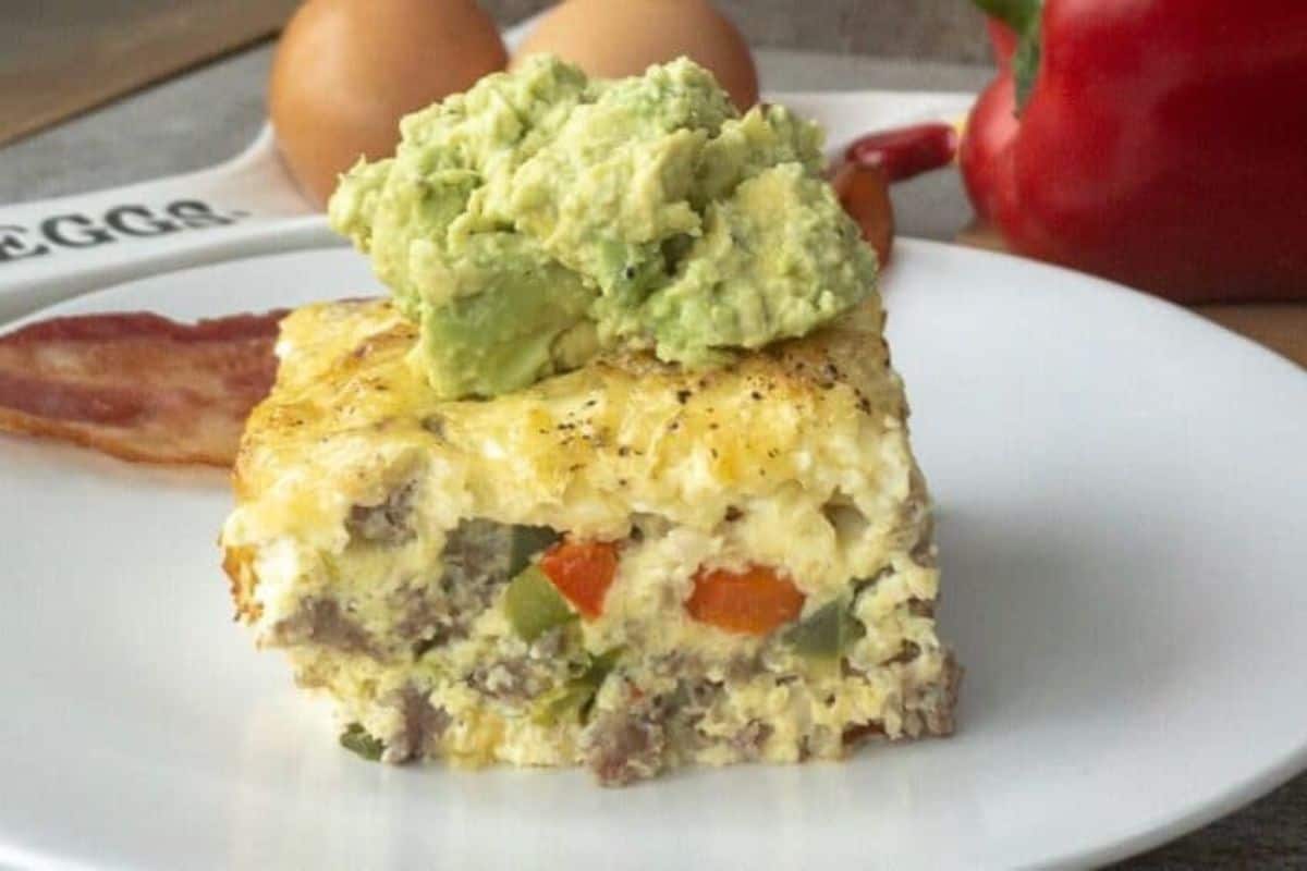 A slice of egg casserole topped with avocado served with bacon on a white plate.