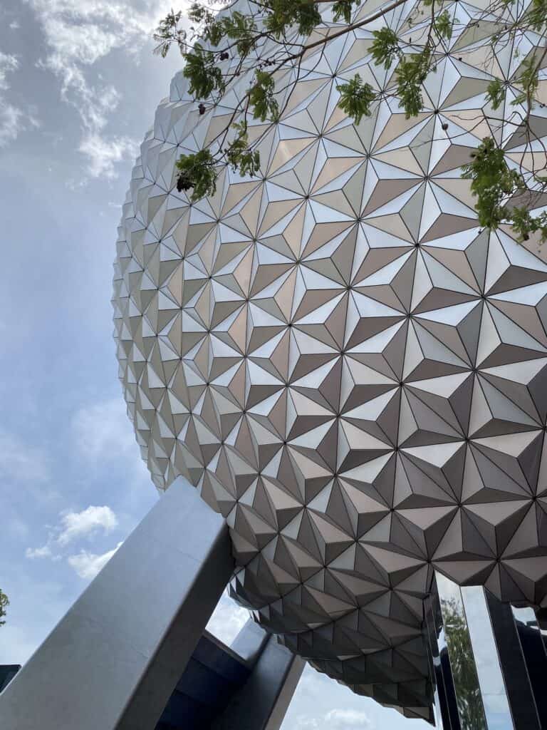 Geodesic dome with a textured white surface supported by large beams, framed against a partly cloudy sky, with foliage emerging from its edges.