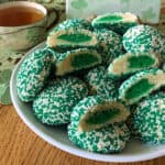 A plate of green and white sprinkled cookies, some cut in half to show green centers, next to a cup of tea on a table with a lacy placemat.