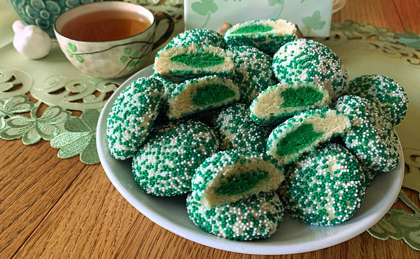 A plate of green and white sprinkled cookies, some cut in half to show green centers, next to a cup of tea on a table with a lacy placemat.