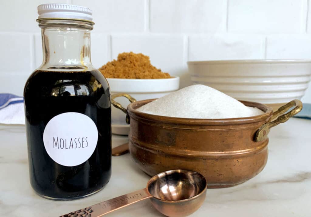A glass bottle of molasses alongside bowls of brown sugar and white sugar with a copper measuring spoon.