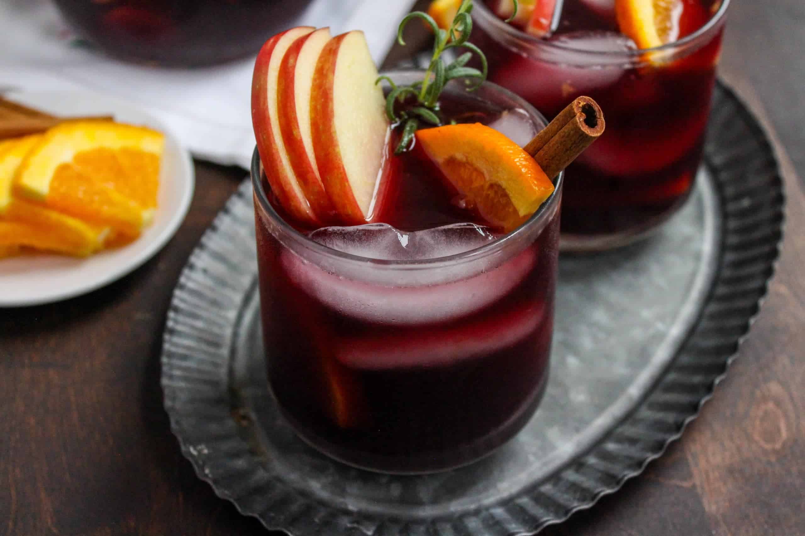 A glass of red wine bourbon sangria with apple slices and cinnamon sticks sitting on a metal plate.