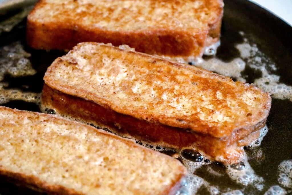 Three slices of french toast cooking in a skillet, sizzling in melted butter and turning golden brown.
