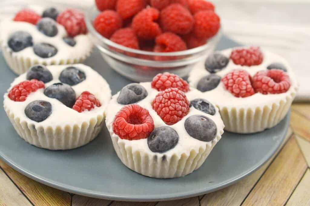 A plate of mini cheesecakes topped with fresh raspberries and blueberries on a wooden surface.