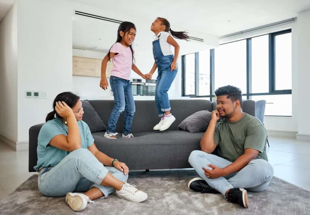 Two children happily jumping on a couch, while a man and woman sit on the floor, looking stressed.