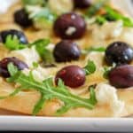 A close-up of a goat cheese flatbreads topped with caramelized onions, arugula, and black blueberries, served on a white plate.