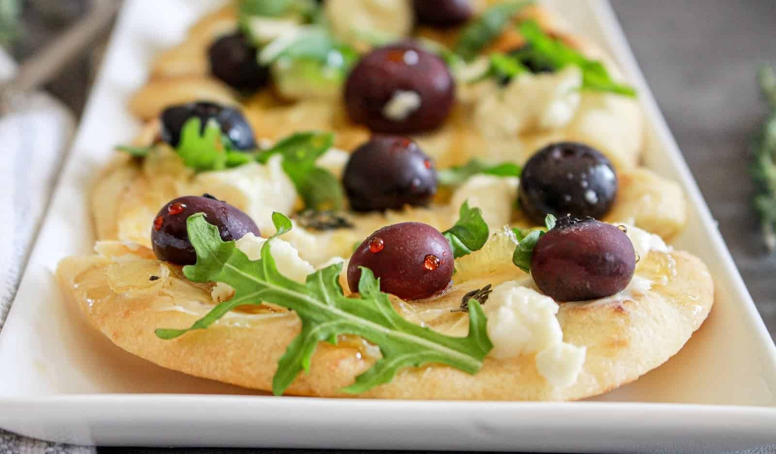 A close-up of a goat cheese flatbreads topped with caramelized onions, arugula, and black blueberries, served on a white plate.
