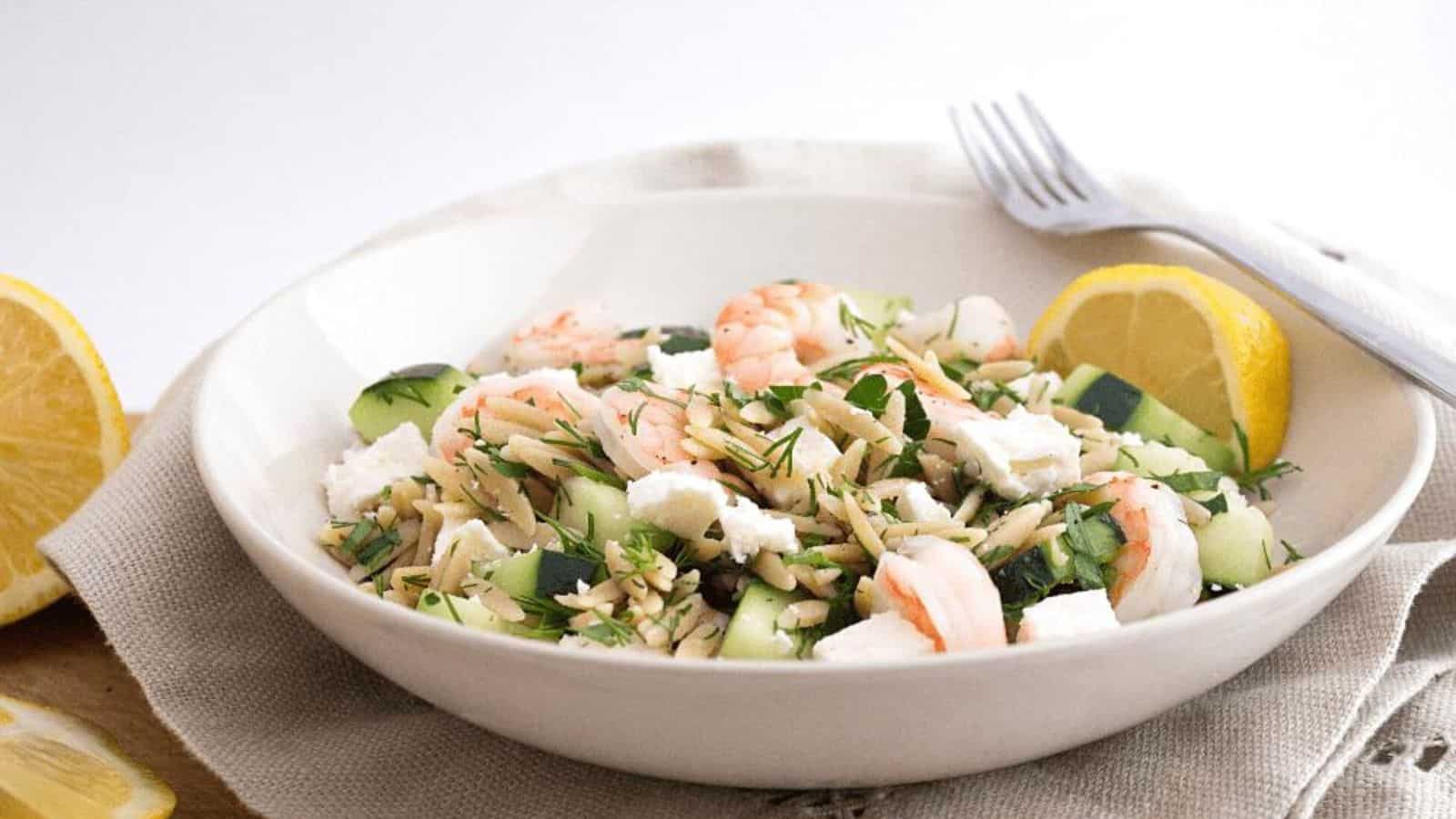 A shallow white bowl with orzo, shrimp, sliced cucumbers, and feta cheese.