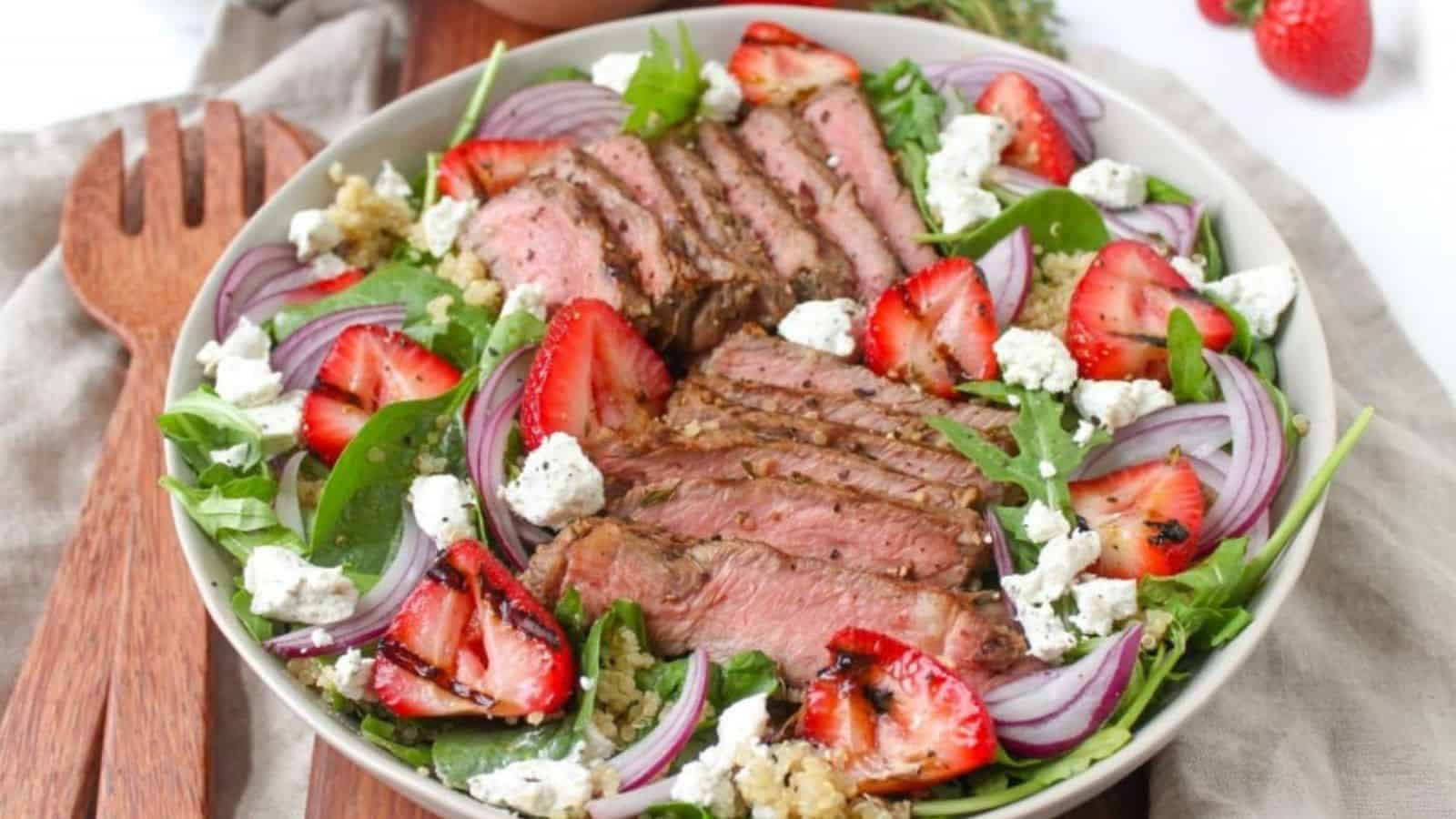 A shallow bowl with greens, grilled steak, strawberries, and crumbled goat cheese.
