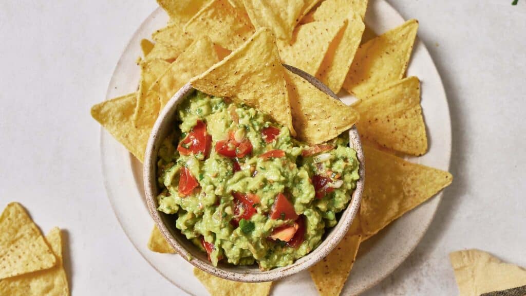 A bowl of guacamole surrounded by tortilla chips on a light surface.