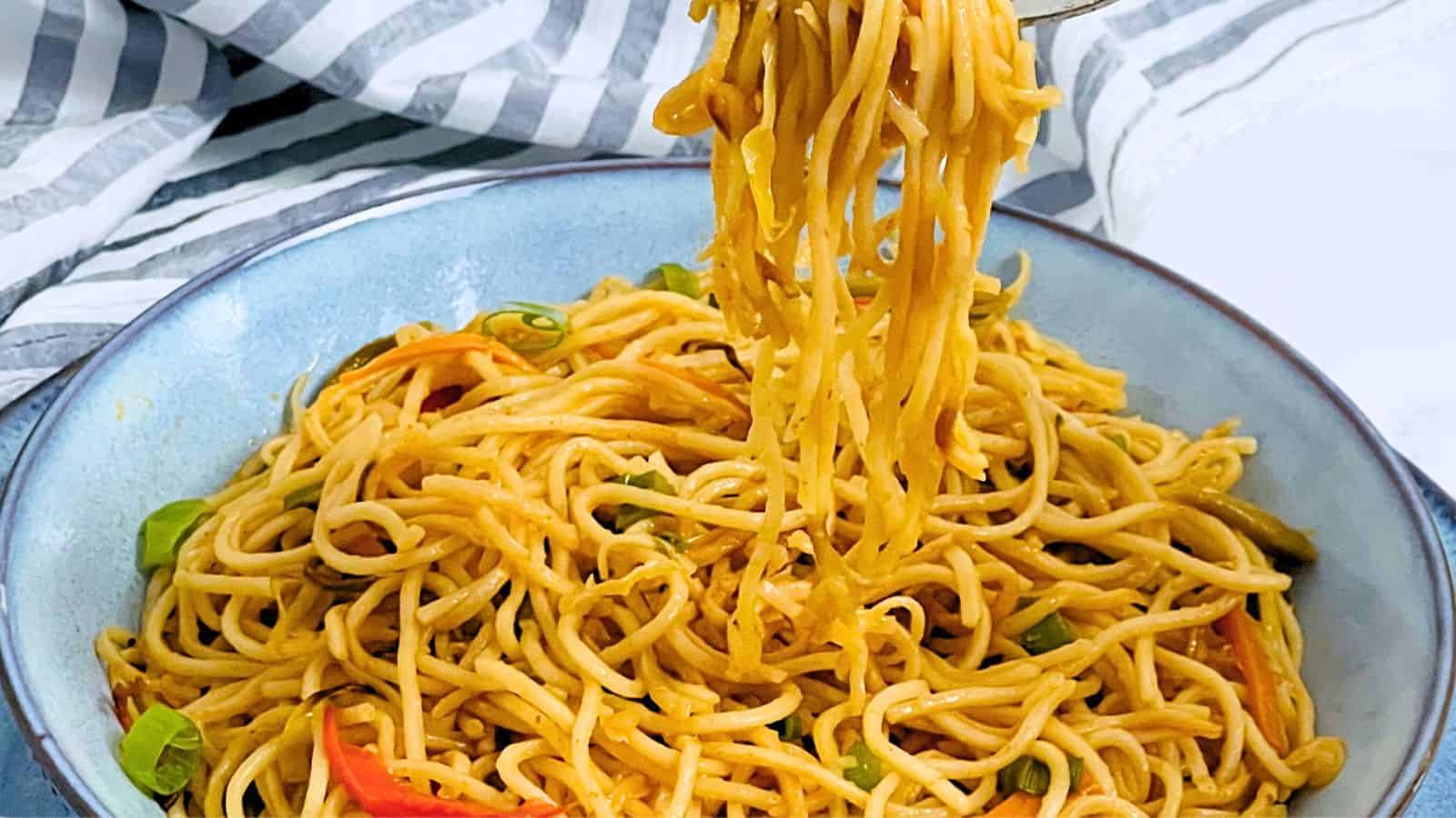 A bowl of Hakka Noodles with vegetables, a fork lifting some noodles, placed on a blue dish with a striped napkin on the side.