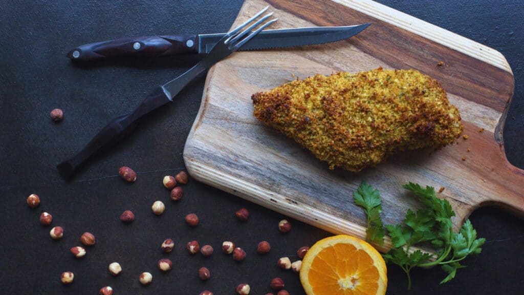 A breaded turkey breast on a wooden cutting board, with a knife and fork beside it, surrounded by hazelnuts and a slice of orange on a dark surface.