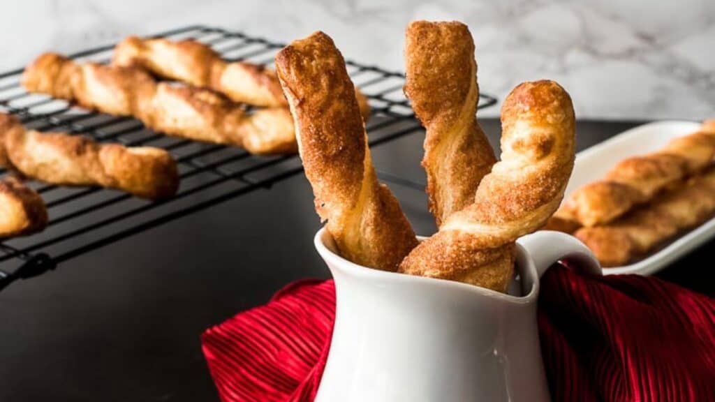 Freshly homemade cinnamon twists in a white cup with more on a cooling rack in the background.