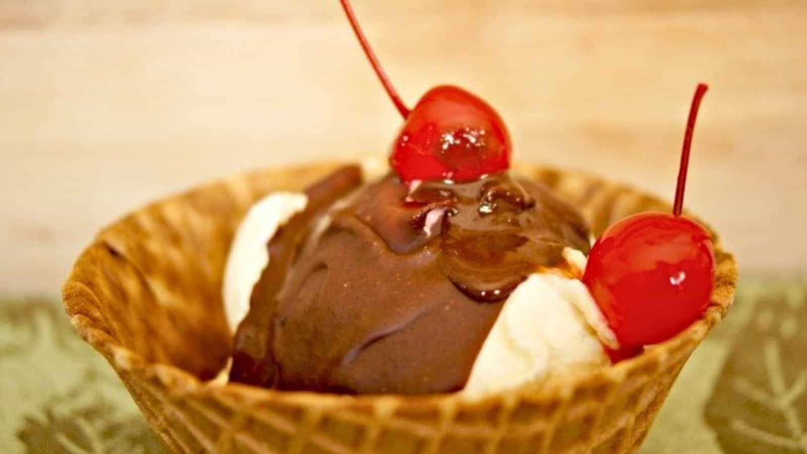 Image shows A waffle cone bowl containing vanilla ice cream topped with magic shell and two cherries.