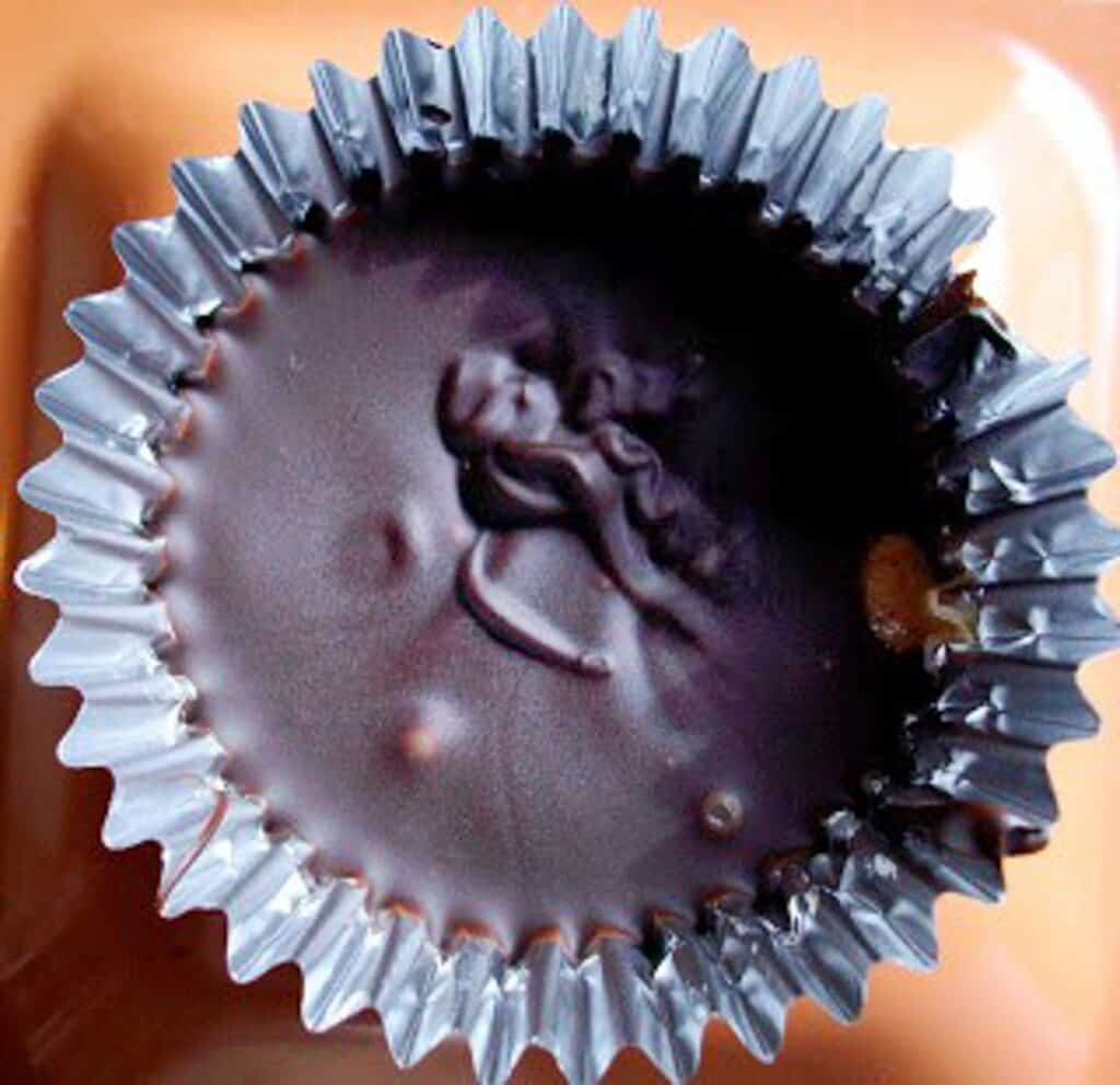 A close-up view of a dark chocolate bonbon in a silver foil wrapper, showing a glossy surface with a slight reflection and a few crumbs.