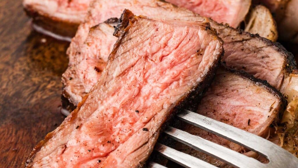 Close-up image of sliced, medium-rare blackened steak on a wooden board, with a fork resting beside the slices.